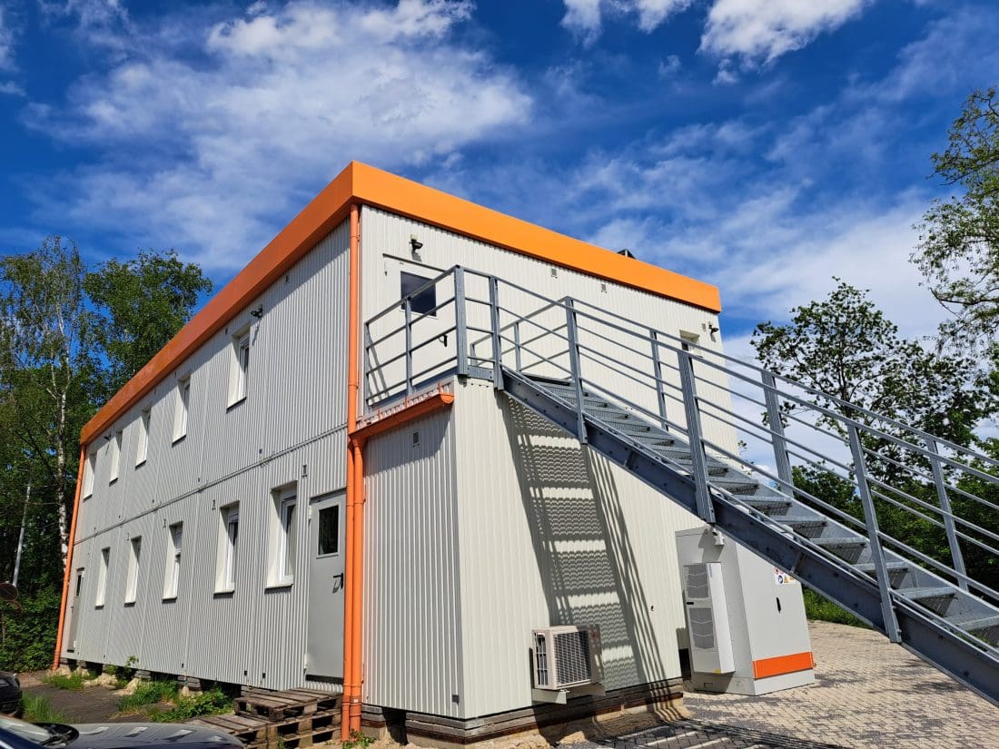 Ein zweistöckiges modulares Gebäude mit orangefarbenen Verzierungen und einer Außentreppe unter einem teilweise bewölkten Himmel.