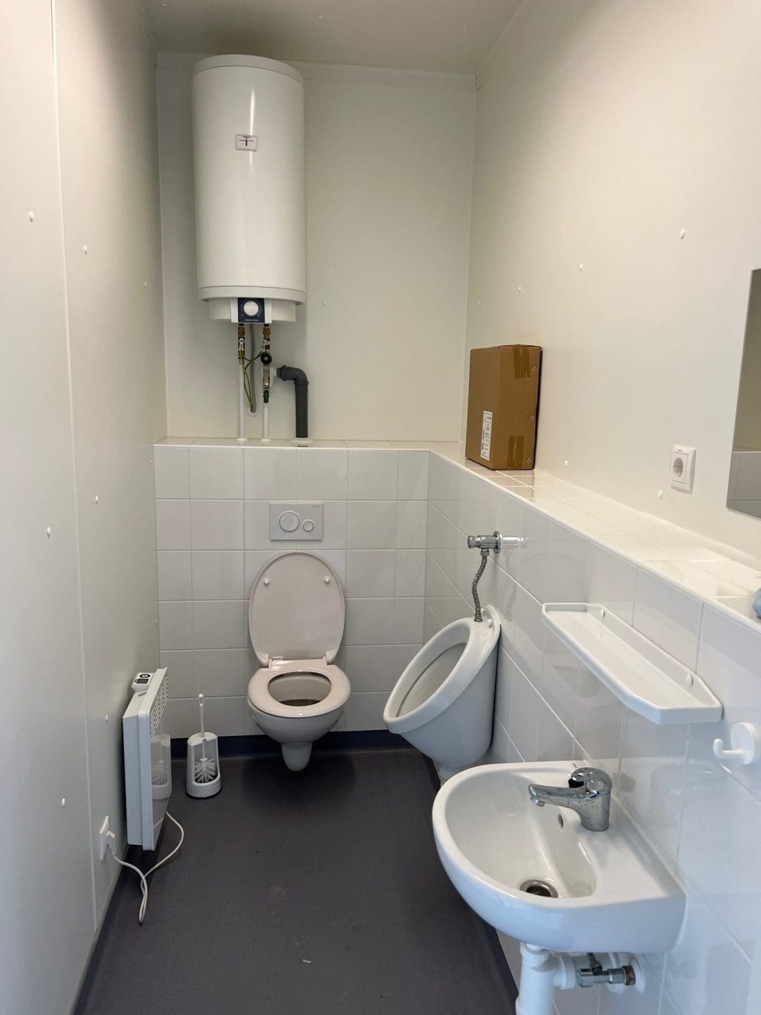 Ein kleines Badezimmer mit Toilette, Urinal, Waschbecken, wandmontiertem Warmwasserbereiter und diversen Einrichtungsgegenständen wie Heizkörper, Seifenschale und Toilettenbürste. Auf dem gefliesten Wandregal steht ein Karton.